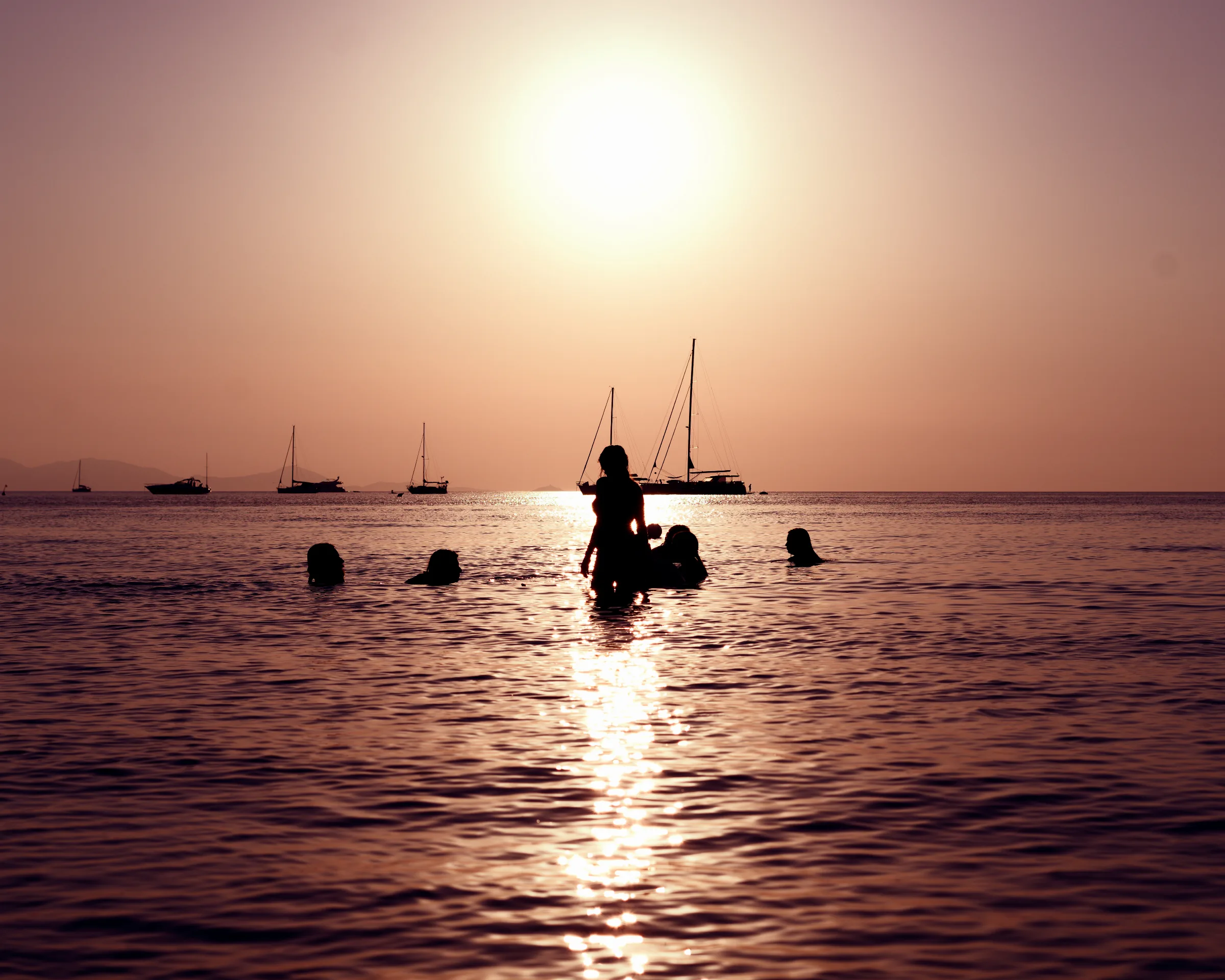 Una persona in silhouette cammina nell’acqua bassa al tramonto; il sole si riflette sul mare e alcune barche a vela sono all’orizzonte.