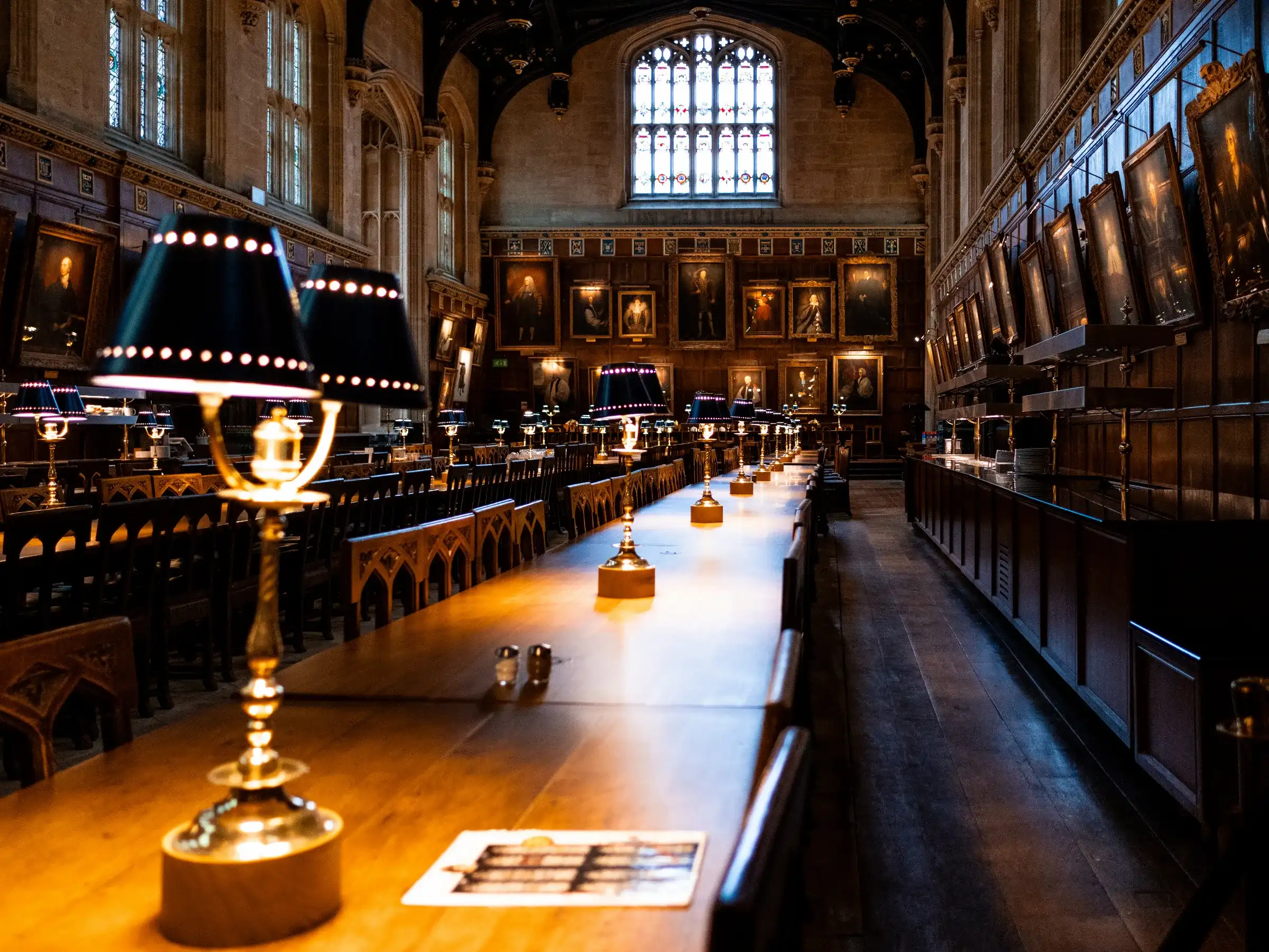 La Sala Grande Christ Church a Oxford, con lunghi tavoli di legno illuminati da lampade, ritratti alle pareti e una grande vetrata sul fondo.