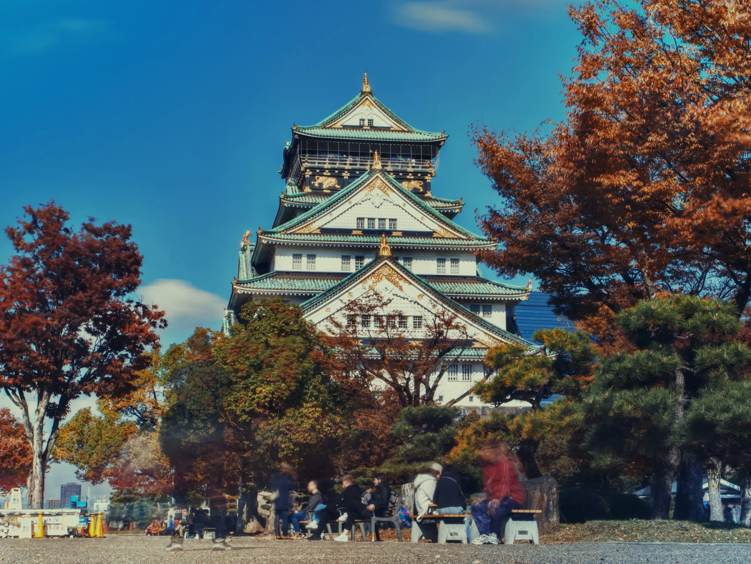 Il castello di Osaka si staglia al centro, circondato da alberi con foglie autunnali; alcune persone camminano sul piazzale in primo piano.