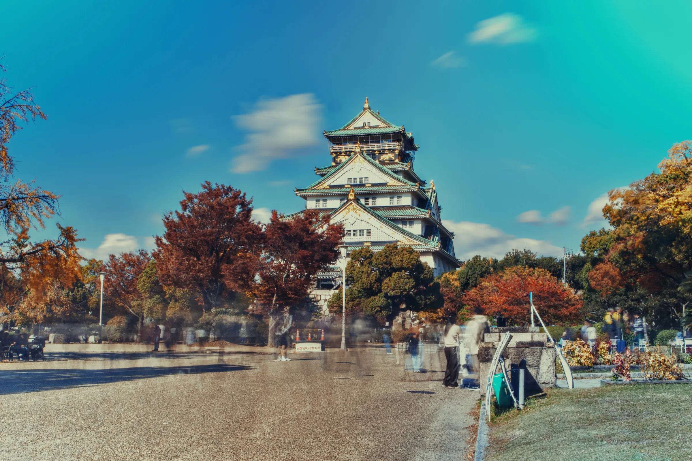 Il Castello di Osaka fotografato con slow shutter: il castello a più livelli si staglia tra alberi dai colori autunnali, con persone mosse in primo piano e cielo azzurro.